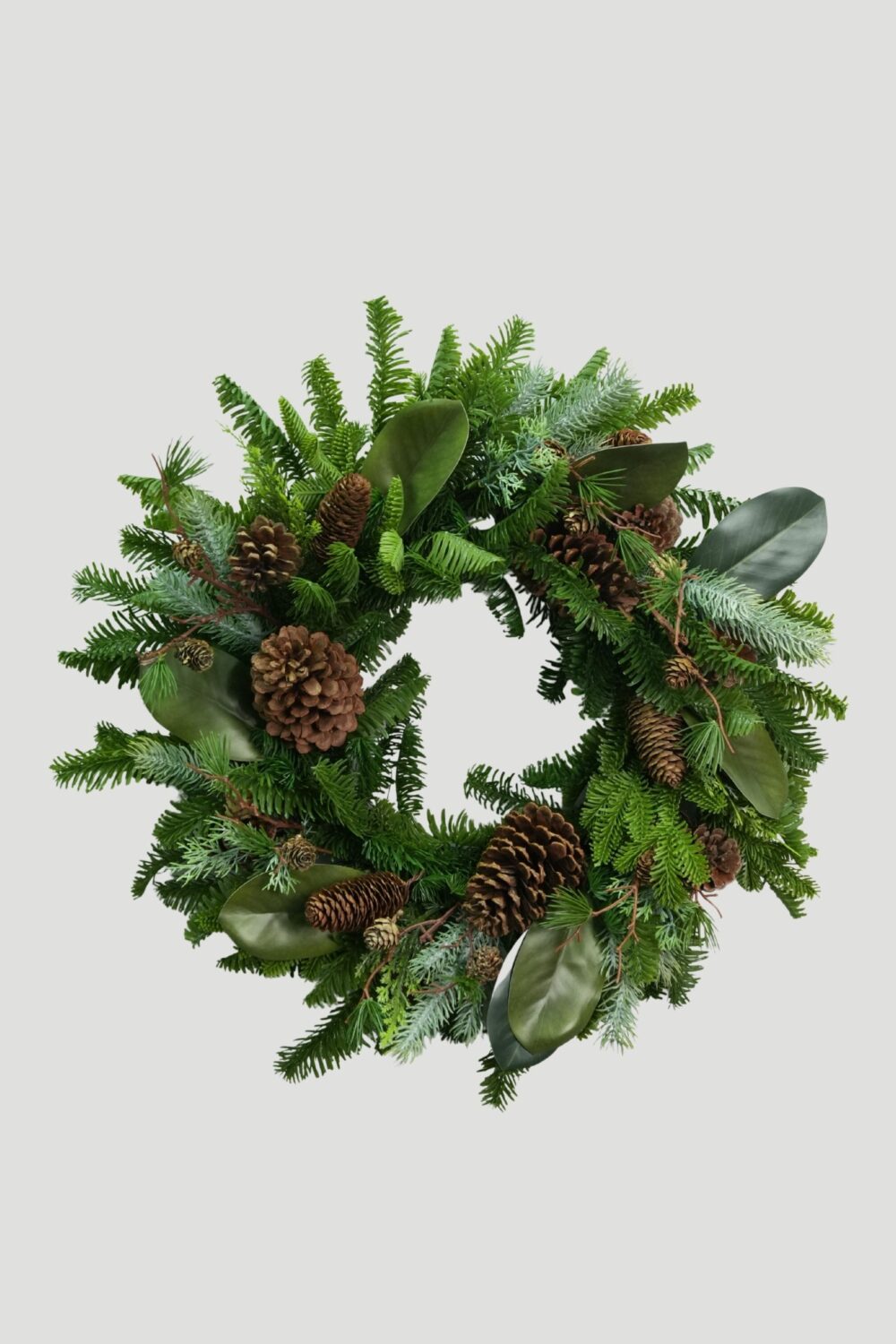 Full view of a 56cm Feel Real Mixed Evergreen Wreath with Magnolia Leaves and Pinecones against a plain background
